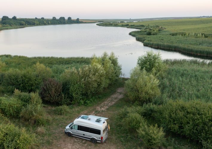 Luftaufnahme vom VW Grand California am ruhigen Seeufer in Rumänien, umgeben von grüner Natur