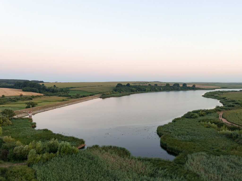 Luftaufnahme des Acumularea-Crestur-Sees in Rumänien mit Damm, Schilfgürtel und Feldern im Abendlicht.