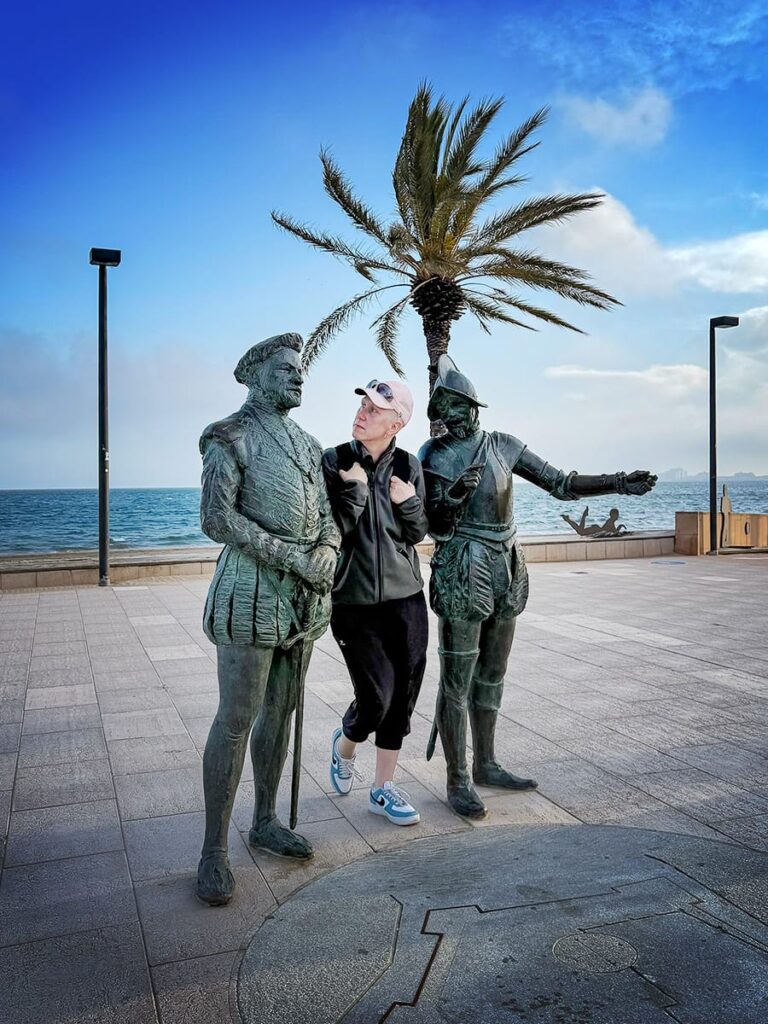 Person steht fragend zwischen zwei Bronzefiguren an der Promenade von Roses mit Meer und Palme im Hintergrund.