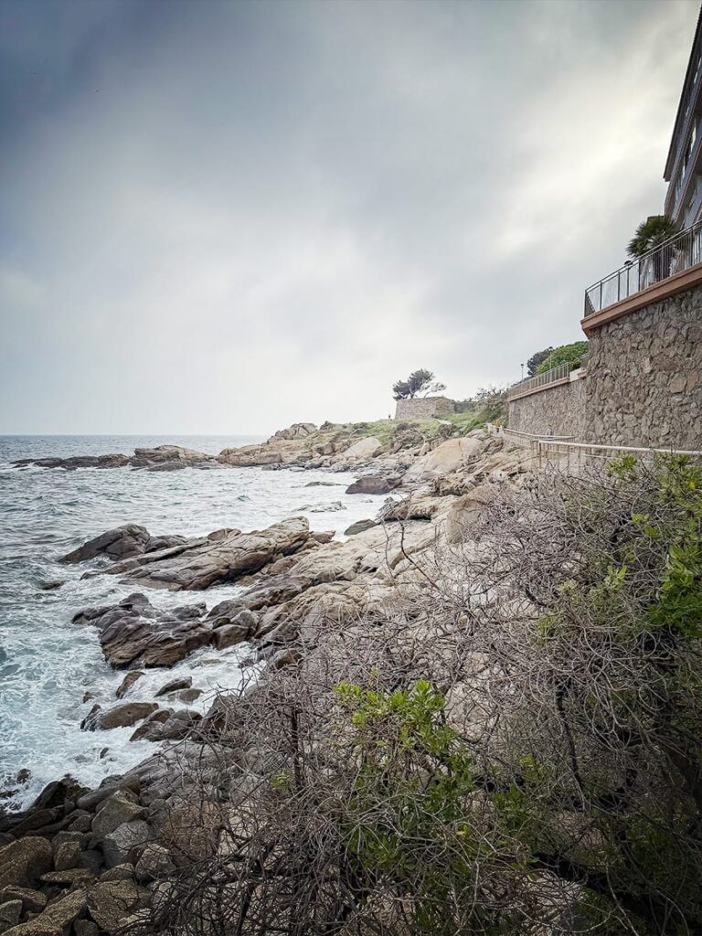 Felsige Küste bei Roses mit Fußweg zwischen Meer und Häusern.