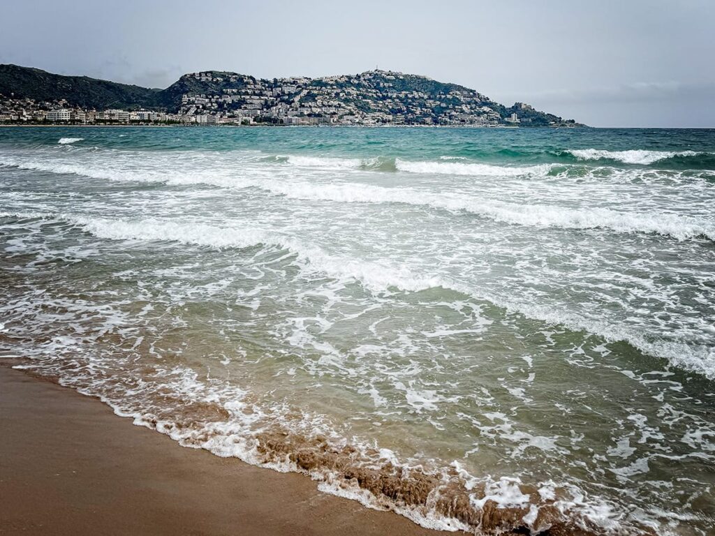 Wellen rollen an den Strand von Roses an der Costa Brava, im Hintergrund die Stadt und Hügel.