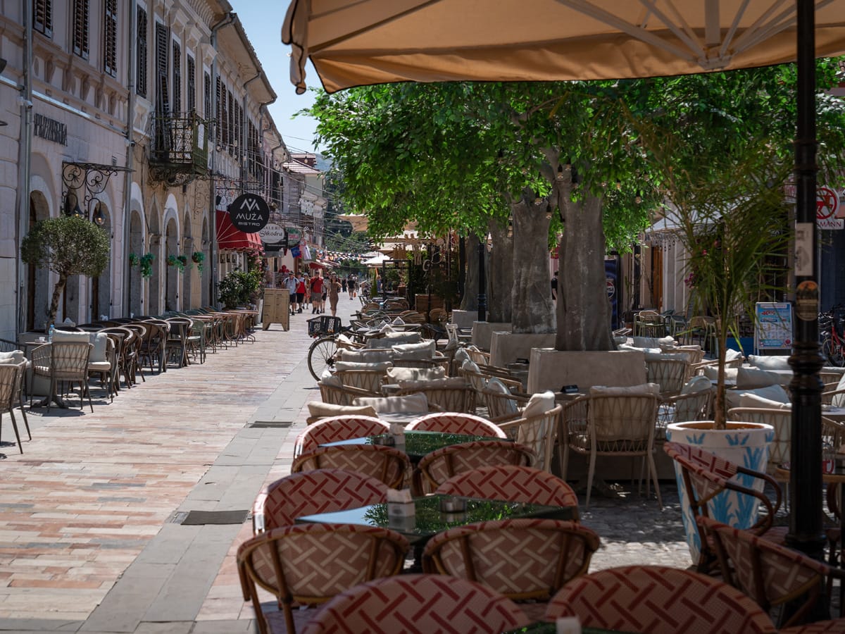 Fußgängerzone Kolë Idromeno in Shkodra mit Cafés, schattenspendenden Bäumen und wenigen Besuchern an einem warmen Nachmittag.