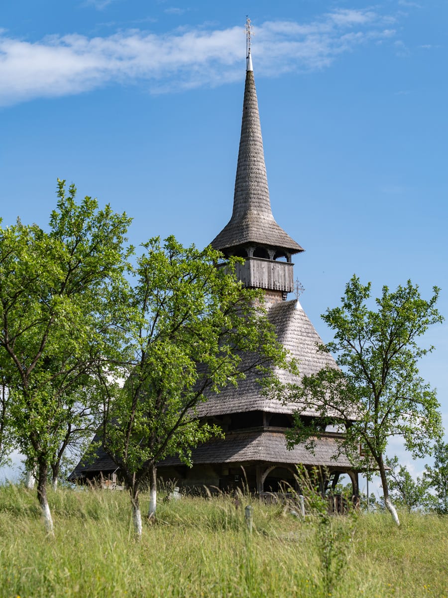 UNESCO-Holzkirche auf dem Jbâr-Hügel in Bârsana, Maramureș, mit ihrem schlanken Holzturm zwischen grünen Bäumen.