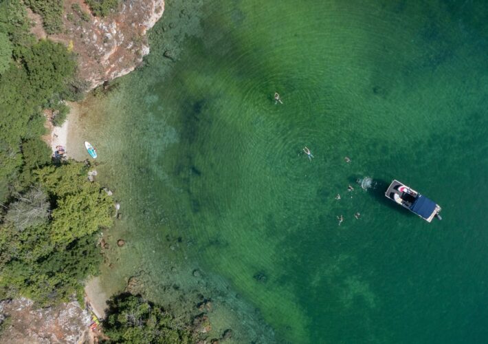 Drohnenaufnahme einer kleinen Badebucht am Südufer des Ohridsees mit Ausflugsboot, Badenden und smaragdgrünem Wasser.