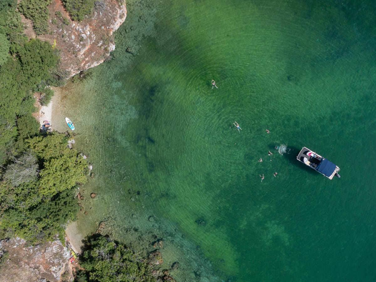 Drohnenaufnahme einer kleinen Badebucht am Südufer des Ohridsees mit Ausflugsboot, Badenden und smaragdgrünem Wasser.