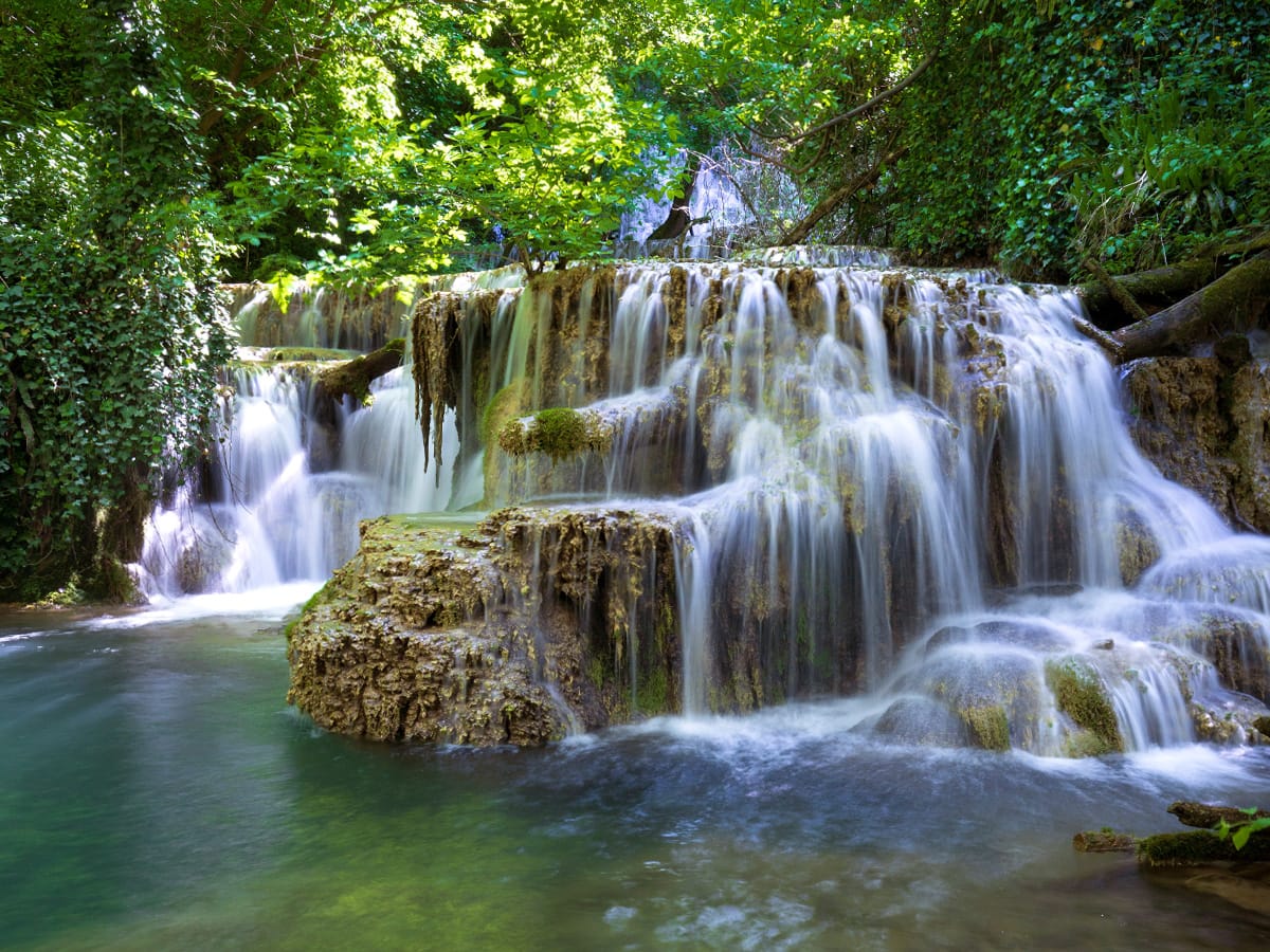 Krushuna-Wasserfälle mit moosbewachsenen Kalkstufen und einem türkisgrünen Wasserbecken unter dichtem Blätterdach.