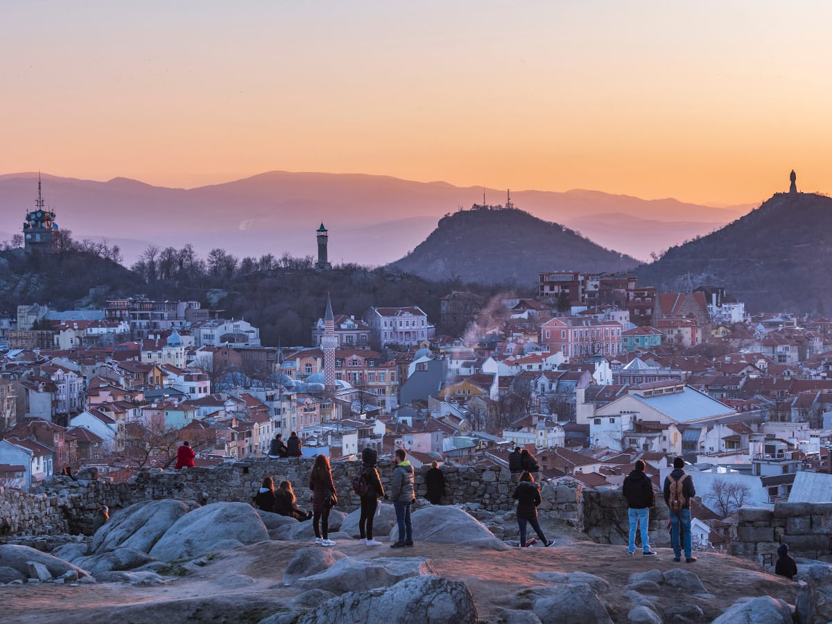 Menschen auf einem felsigen Aussichtspunkt über Plowdiw, dahinter Dächer, Stadthügel und ein orangefarbener Abendhimmel.